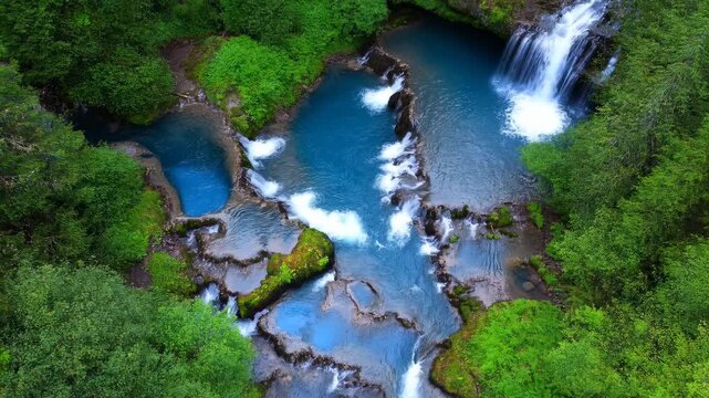 High angle view of terraced turquoise waterfalls flowing through dense temperate rainforest, with serpentine channels, foamy plunge pools, wet dark rock, and rich green canopy