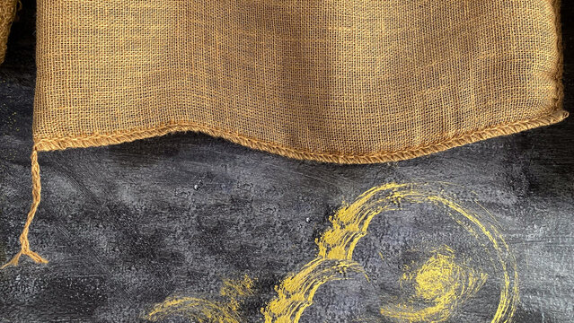 Textured brown burlap sack cloth partially covering a dark grunge background with golden brush strokes. Top view conveying an earthy raw aesthetic, representing creative rustic packaging copy space.