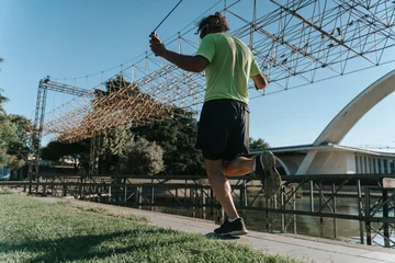 man running outdoors on a bridge with water body in the background, low angle view, active lifestyle, green and black clothing, metal structure, natural setting, clean environment. © uladzimirzuyeu