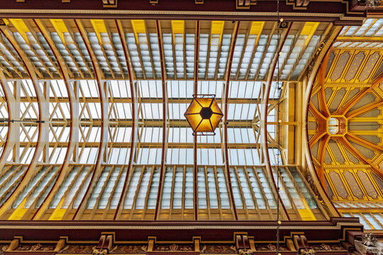View of the ornate Victorian glass and iron roof of Leadenhall Market with intricate yellow and red painted details and a central hanging lantern in London, England, United Kingdom.