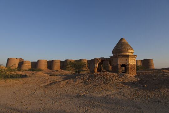 View of the ancient Derawar Fort with its massive circular bastions and a crumbling stone structure in the foreground under a clear blue sky in Derawar, Punjab, Pakistan.