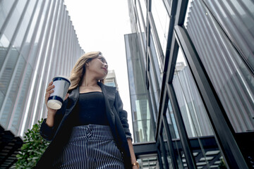 Fototapeta premium Asian businesswoman walks briskly with mouth slightly open and gaze lifted up and right while holding coffee beneath Bangkok's towers, on a busy sidewalk beneath Bangkok's glass skyline.