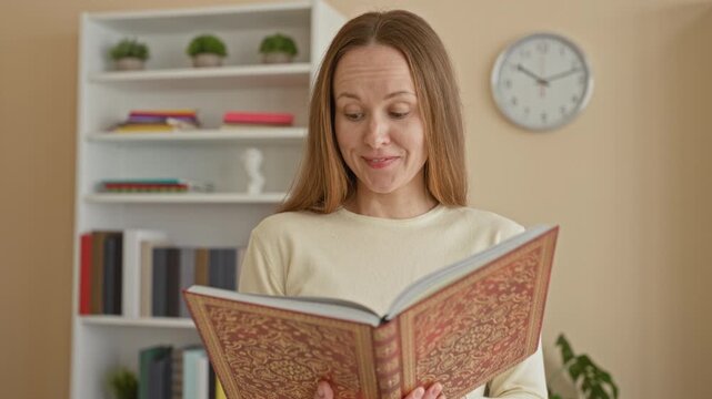 Young caucasian woman reading an ornate hardcover book, hands holding open pages, bookshelf and wall clock visible in studio room with plants and shelves; concentration.