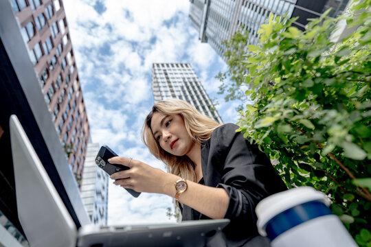 Asian businesswoman with long blonde-dyed wavy hair turns her gaze toward her phone screen, laptop resting on her lap surrounded by dense green foliage in Bangkok.