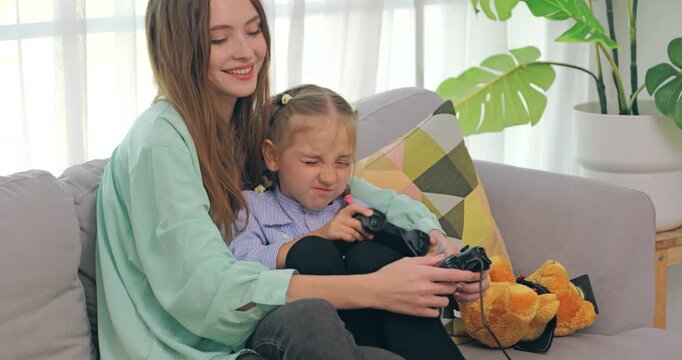 A happy mother and little girl playing video games together with controllers on the living room sofa.