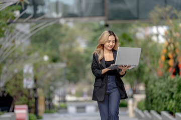 Fototapeta premium Asian businesswoman with long blonde wavy hair strides along a landscaped garden path in Bangkok's business district, holding an open laptop and glancing down toward its glowing screen.