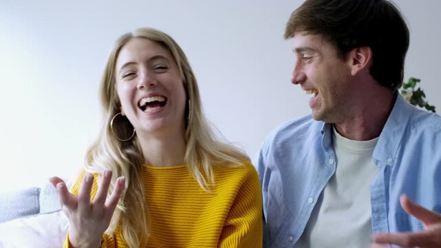 A cheerful young couple smiles and waves, engaged in a lively video call with friends or family from their home. They express happiness and connection while communicating virtually.