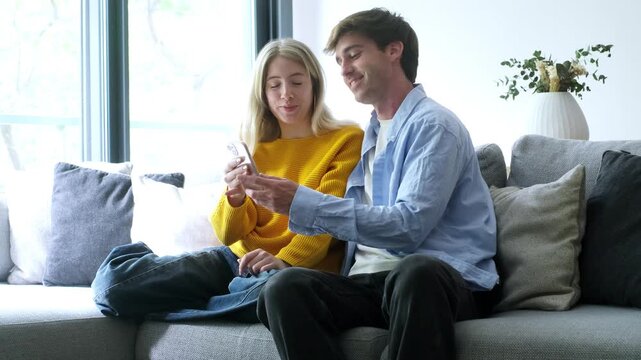 A young couple sits on a couch, looking at a smartphone, smiling and waving, as they enjoy a video call or browse content together at home.