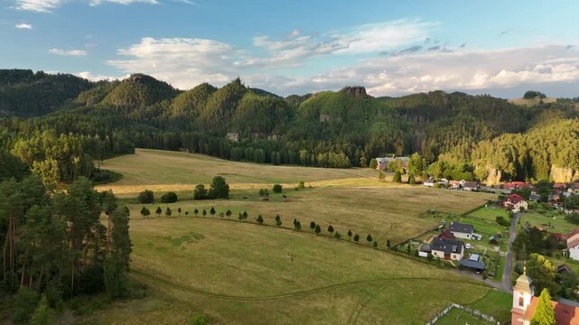 Drone shot of a village with meadows and a chapel below sandstone rock pinnacles at sunset in summer in Czech Republic