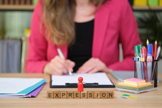 A woman in a pink blazer writes intently in a notebook, surrounded by colorful stationery and notes. The word expression formed with wooden blocks sits prominently on the desk.