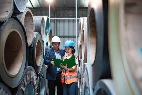Factory manager and female engineer reviewing documents while walking between steel coils, checking inventory, material stock, quality control and manufacturing operations.