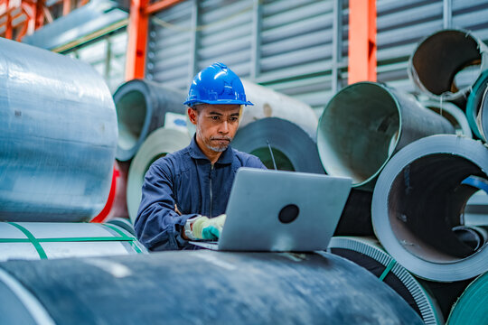 Industrial worker working on laptop among steel coils in factory warehouse, checking inventory records, production data, material stock and manufacturing operations.