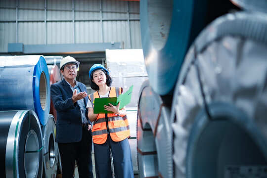 Factory manager and female engineer checking steel coil inventory in warehouse, discussing material stock, quality control, production planning and industrial operations.