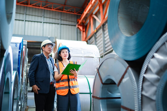 Factory manager and female engineer inspecting steel coil inventory in warehouse, reviewing material stock, quality control, production planning and manufacturing operations.