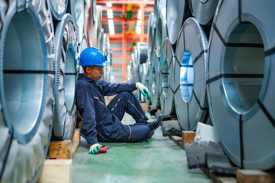 Factory worker resting between steel coils after inspection in industrial warehouse, showing physical work, fatigue, workplace safety and manufacturing occupation.