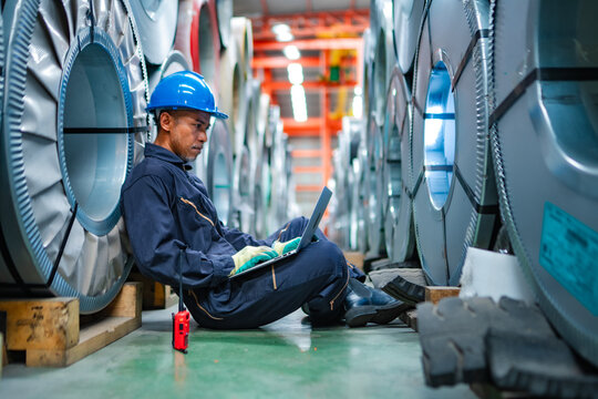 Industrial worker using laptop while sitting between steel coils in factory warehouse, checking inventory data, production records, material stock and manufacturing operations.