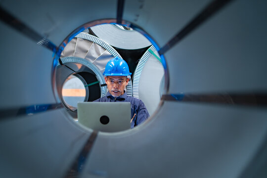 Factory worker using laptop viewed through center of steel coil, checking inventory records, production data and material management in industrial warehouse.