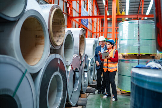 Female engineer using tablet with factory manager among steel coils, checking inventory, material stock, production planning and warehouse operations.