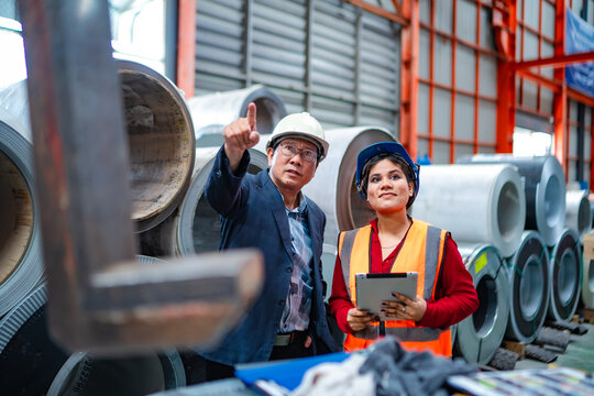 Factory manager pointing toward steel coil storage while female engineer holds tablet, discussing inventory, warehouse inspection, production planning and industrial operations.