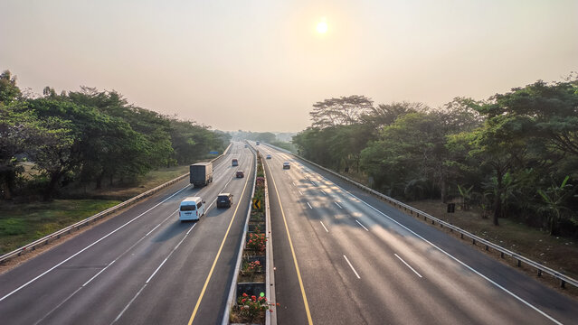 A divided highway stretching through a green tropical landscape during a warm hazy morning sunrise, with vehicles traveling along the road and trees lining both sides