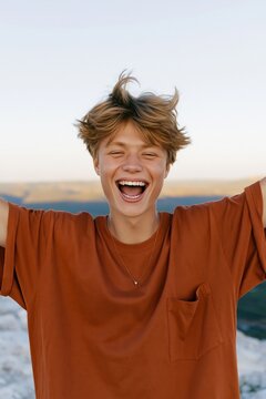 Laughing teenage boy with tousled hair and raised arms on a cliff for joyful outdoor background and youth lifestyle design