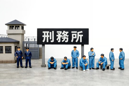 刑務所の看板の前で青い作業着を着てしゃがんだり立ち話したりする受刑者と看守のミニチュア