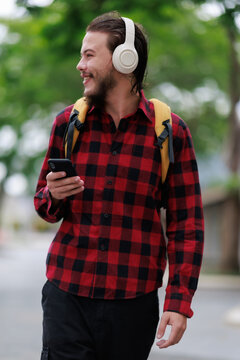 Young man wearing a plaid shirt, backpack, and wireless headphones walking outdoors, looking at his smartphone while smiling and enjoying his favorite music