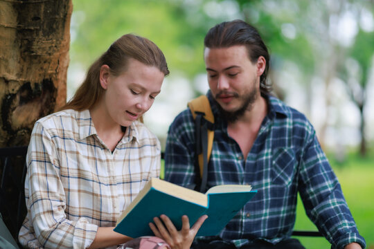 Young student couple learning outdoors, reading an open book together in a park, sharing knowledge and cooperating for education, focusing on academic study