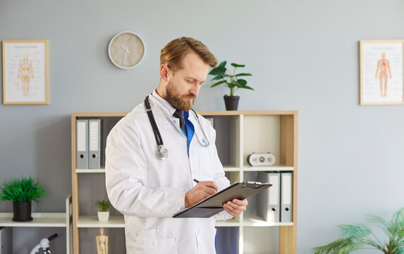 Male physician or general practitioner writing down notes, holding clipboard, posing in medical office, has serious expression, wearing white coat with stethoscope, preparing for appointment