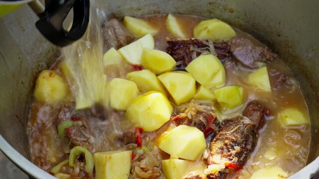 Traditional stew simmering in a large cast-iron kazan cauldron outdoors. Hot steam and rich ingredients highlight authentic ethnic cuisine