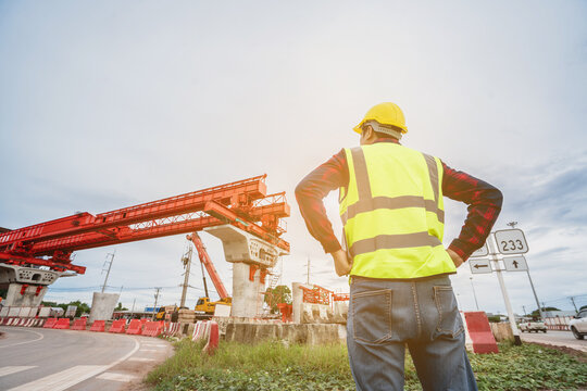 Construction foreman and engineer, wearing safety equipment, are looking at blueprints at a high bridge construction site where large cranes are operating.