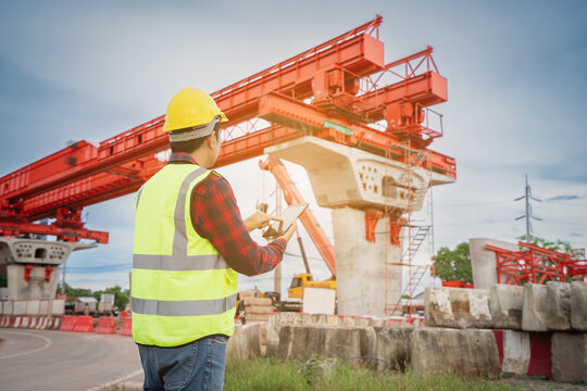Construction foreman and engineer, wearing safety equipment, are looking at blueprints at a high bridge construction site where large cranes are operating.