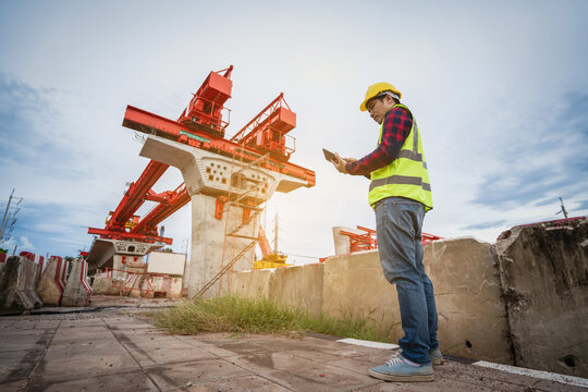 Construction foreman and engineer, wearing safety equipment, are looking at blueprints at a high bridge construction site where large cranes are operating.