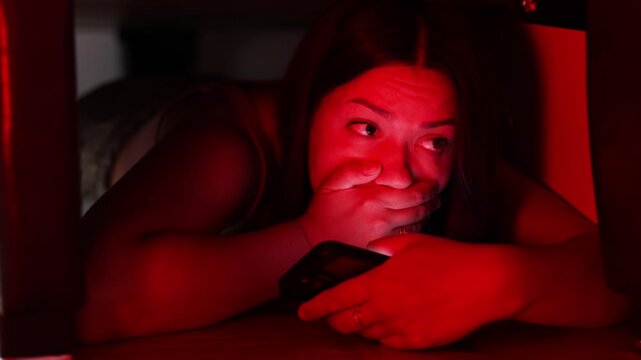 Female criminal hides beneath bed from police. She covers her mouth while using smartphone to contact accomplices.
