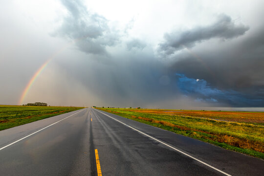 View of a long asphalt road stretching through open fields under a dramatic sky with dark storm clouds and a vibrant rainbow appearing after a rain shower.