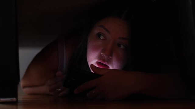 Female criminal hides beneath bed and watches for police. Smartphone keeps her connected with accomplices while emergency lights reveal her face.