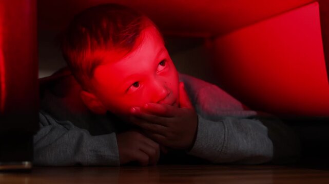 Young boy hides beneath bed after making false police call. Emergency lights illuminate his face while he waits for officers to leave.