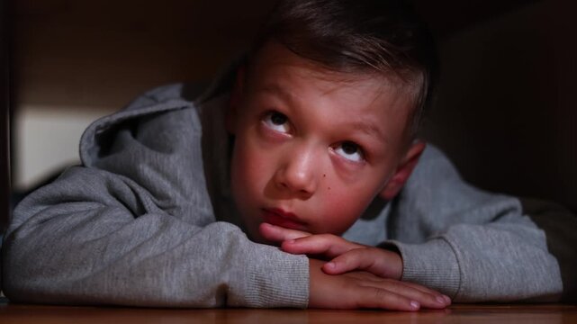 Young boy hides beneath bed in intense fear. Police lights illuminate his face as he opens his mouth in shock.