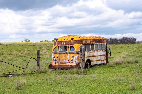 View of an abandoned and rusted yellow school bus sitting in a grassy field under a cloudy sky with a barbed wire fence in the foreground.