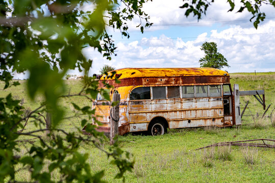 View of an abandoned, rusted yellow school bus sitting in a grassy field under a cloudy sky.