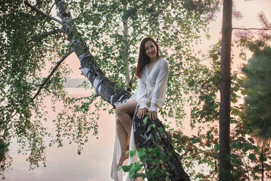 woman is perched on a branch of a tree by a lake at sunset. scene shows green leaves and gentle water. sky has warm colors as the day ends