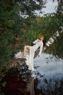 woman in a white dress relaxes on a tree branch over a lake. She sits calmly with her legs hanging near the water, surrounded by green leaves and trees in the afternoon light