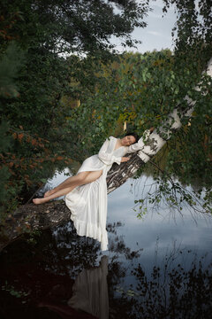 woman lies on a tree branch over a body of water in a forest. She appears relaxed and comfortable as she enjoys the natural surroundings. time is during daylight