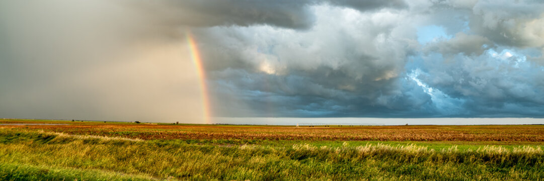 View of a vibrant rainbow arching over a vast agricultural field beneath dramatic, dark storm clouds.