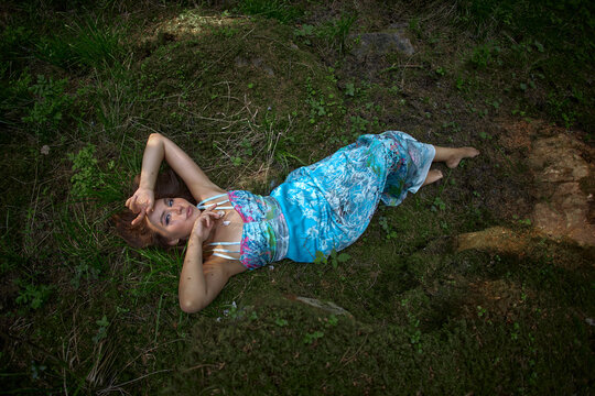 woman rests on the grass wearing a colorful dress. She has her arms raised and appears relaxed. Trees and plants surround her in a natural outdoor setting during the day