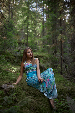 woman rests on the soft moss in a lush green forest. She wears a long dress and looks relaxed amid tall trees during the day. Sunlight filters through the leaves above