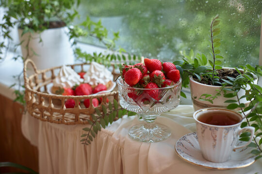 On a table near a window, a crystal bowl holds ripe strawberries. cup of tea sits beside it. Plants add greenery and create a cozy setting. sunlight shines through the glass