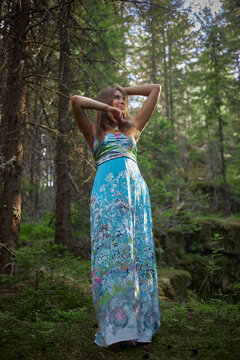 woman stands in a forest, wearing a long floral dress. She poses with her arms raised, surrounded by trees and greenery during a sunny day. light filters through the leaves