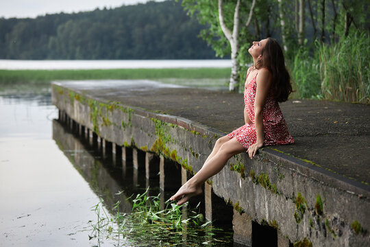 woman in a red dress sits on a wooden dock by a lake. She enjoys the sun and lets her feet rest in the water. Green trees and plants surround the area