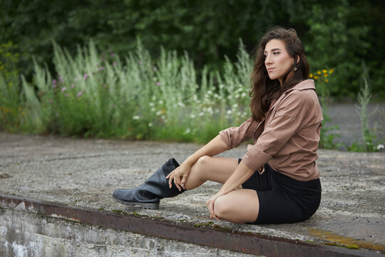 woman rests on a concrete surface in an outdoor area. She wears a brown shirt and a black skirt. Green plants grow around the scene in daylight